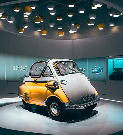 Żółto-biały zabytkowy samochód BMW Isetta na wystawie w muzeum, oświetlony licznymi lampami w kształcie kul.