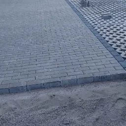 Ułożona szara kostka brukowa przed domem z obrzeżami, widoczne ślady butów w piasku, na palecie pozostałości kostki.