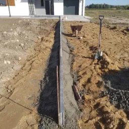 Przygotowanie podłoża pod obrzeże chodnika: wykop, szalunek z prętami zbrojeniowymi, poziomica i sterta tłucznia w pobliżu łopaty i taczki, z widokiem na nowoczesny dom z szarą stolarką.