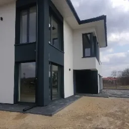 Nowoczesny dom dwukondygnacyjny z płaskim dachem, wykończony w bieli i antracycie, z widocznymi oknami i drzwiami balkonowymi, położony na działce z częściowo ułożoną kostką brukową...