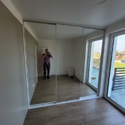 Nowoczesna szafa z przesuwnymi lustrzanymi drzwiami, z odbiciem osoby robiącej zdjęcie, na podłodze panele imitujące drewno, widok przez okno na osiedle domów jednorodzinnych.