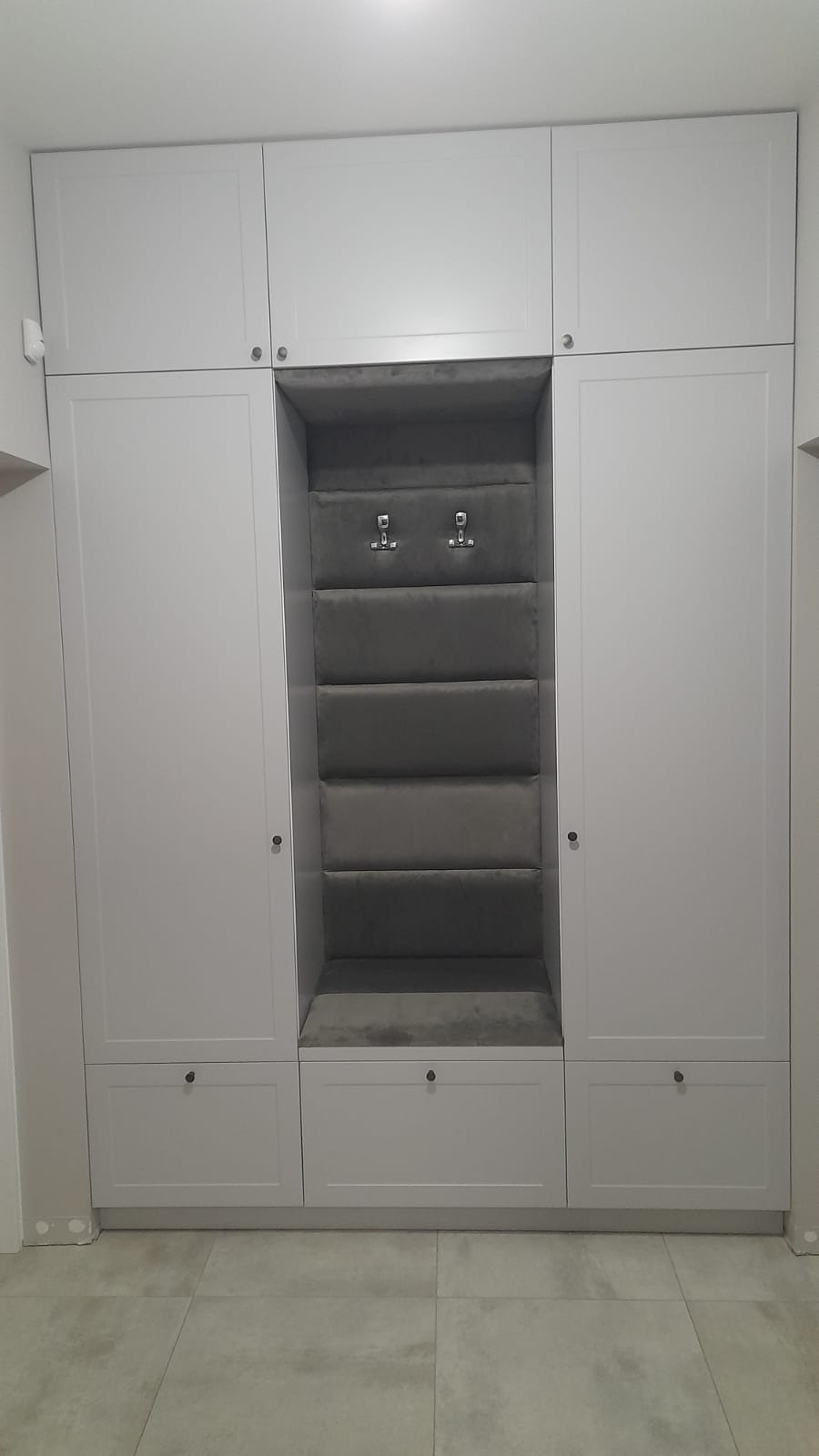 Szafa z wneka tapicerowane i frontami frezowanymi w kolorze jasno szarym.