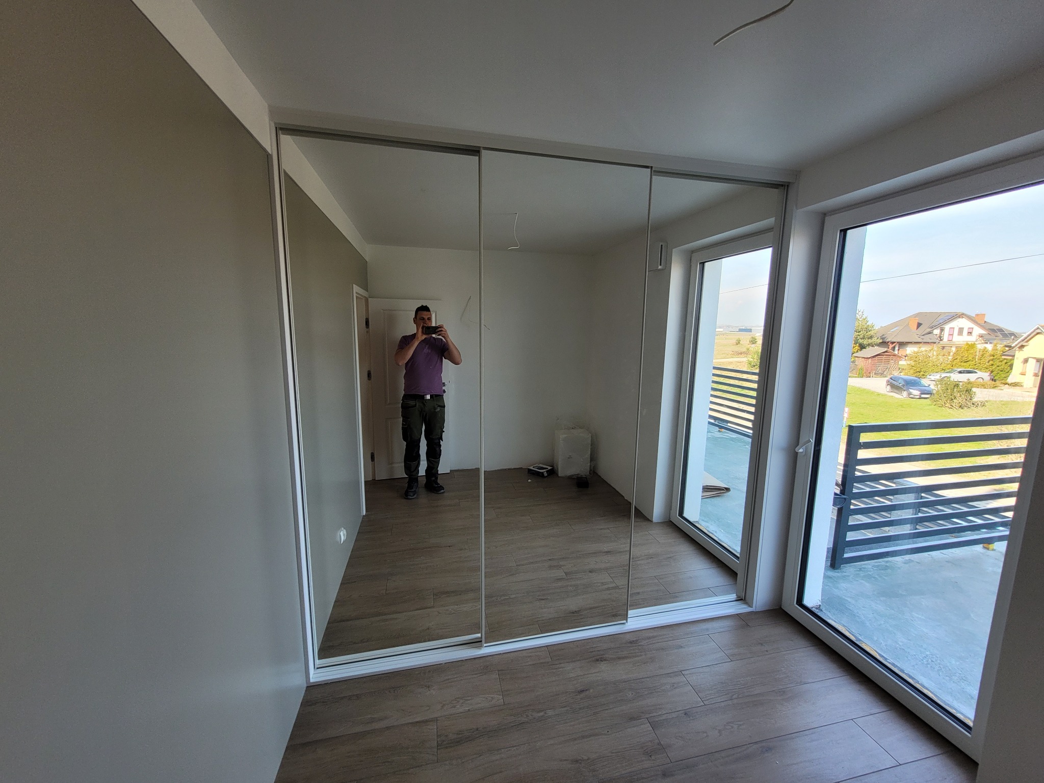 Nowoczesna szafa z przesuwnymi lustrzanymi drzwiami, z odbiciem osoby robiącej zdjęcie, na podłodze panele imitujące drewno, widok przez okno na osiedle domów jednorodzinnych.