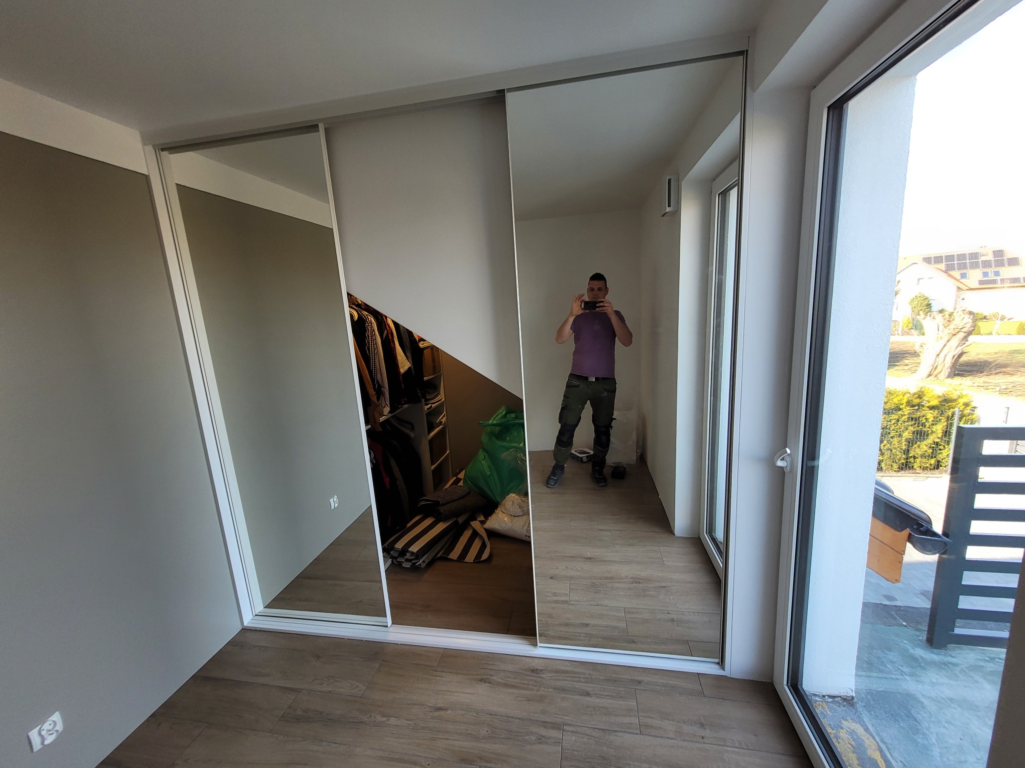 Nowoczesna szafa z przesuwnymi drzwiami lustrzanymi w jasnym wnętrzu, odbicie fotografa robiącego zdjęcie, widoczne ubrania wewnątrz, podłoga z paneli imitujących drewno.