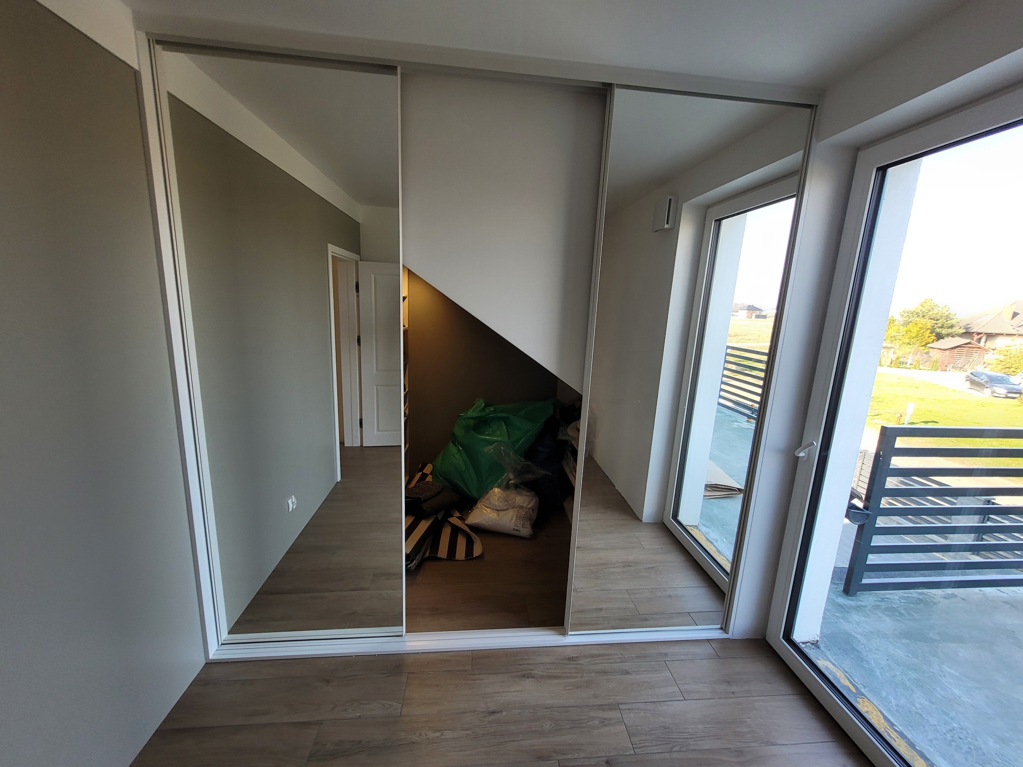 Nowoczesna szafa z przesuwnymi drzwiami lustrzanymi w jasnym wnętrzu, widoczny fragment zagospodarowanego wnętrza za szafą i wyjście na balkon.