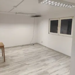 Puste, białe biuro z panelami podłogowymi imitującymi drewno, oświetleniem rastrowym i niedokończonym sufitem podwieszanym.