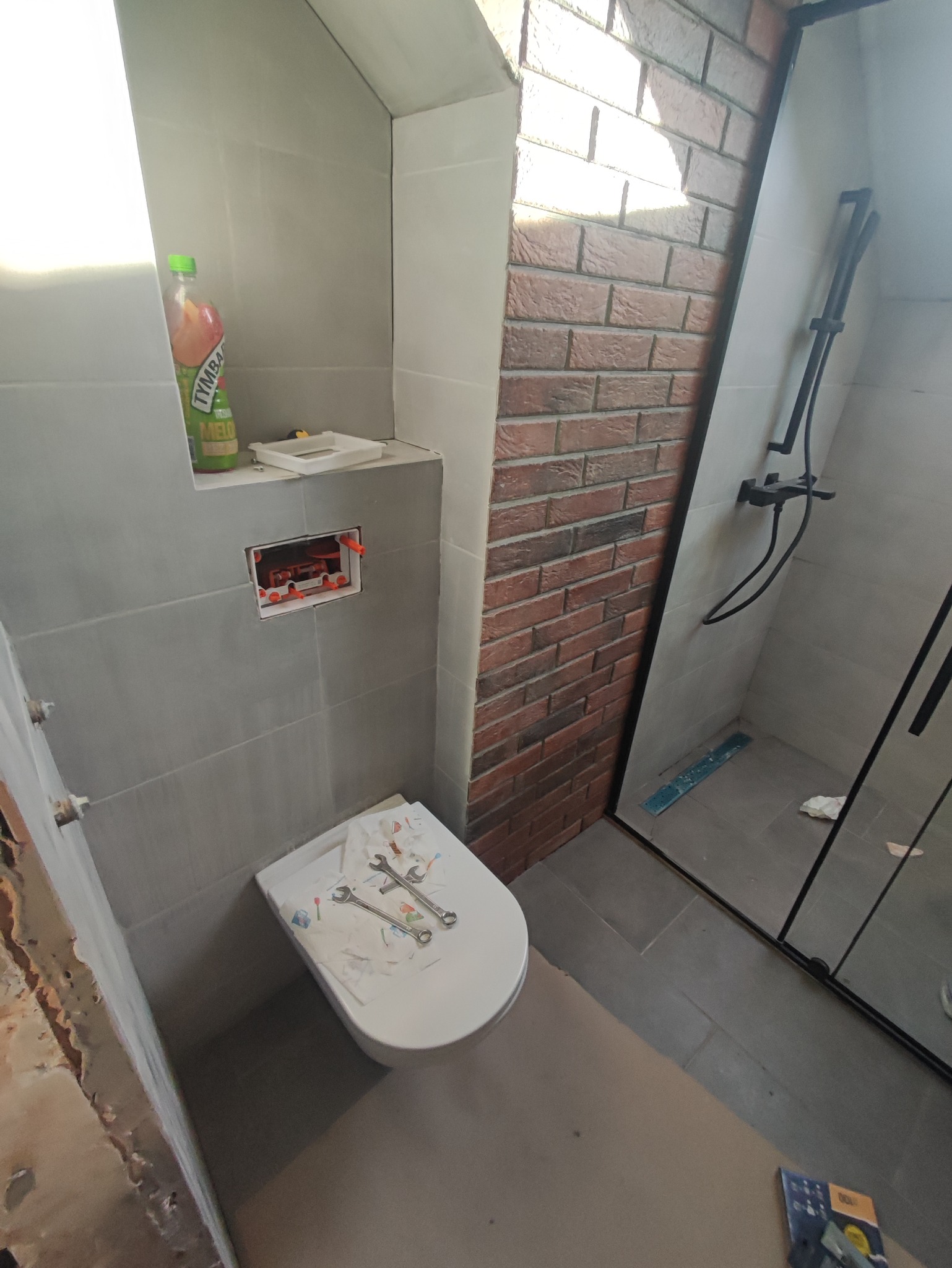 Łazienka w trakcie remontu: częściowo wykończona ściana z cegły, szara glazura, zamontowana toaleta z narzędziami na desce, widoczna kabina prysznicowa.
