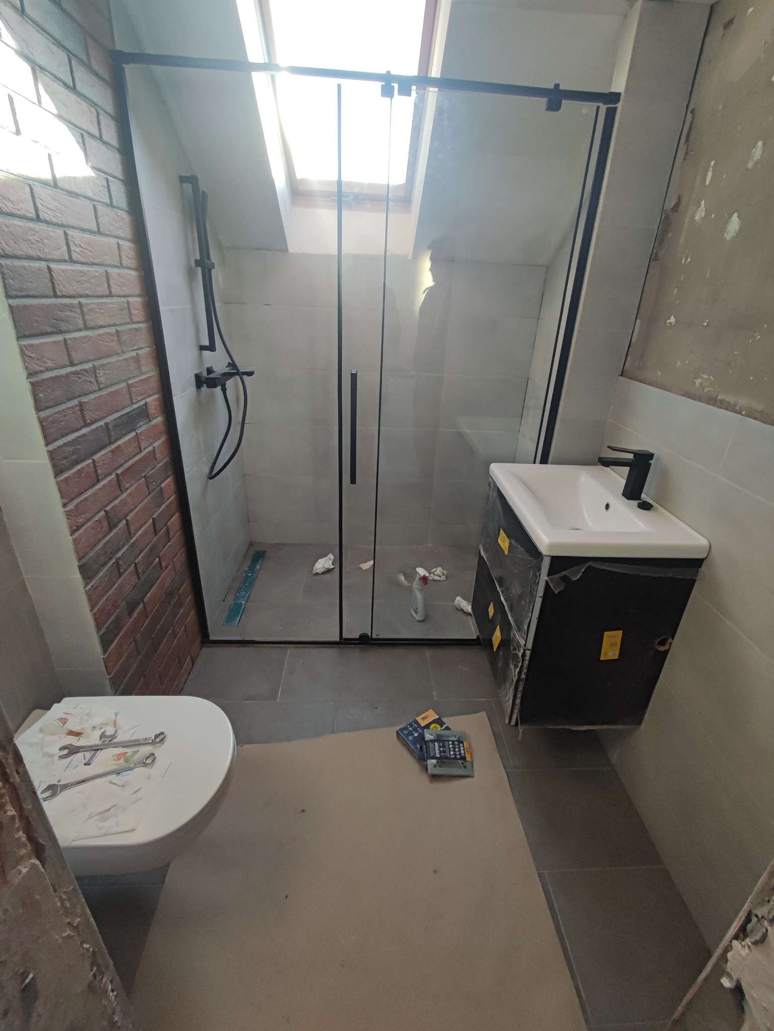Łazienka w trakcie remontu: kabina prysznicowa z czarnymi profilami i armaturą, umywalka z szafką, toaleta z narzędziami, ściana z cegły dekoracyjnej, okno dachowe.