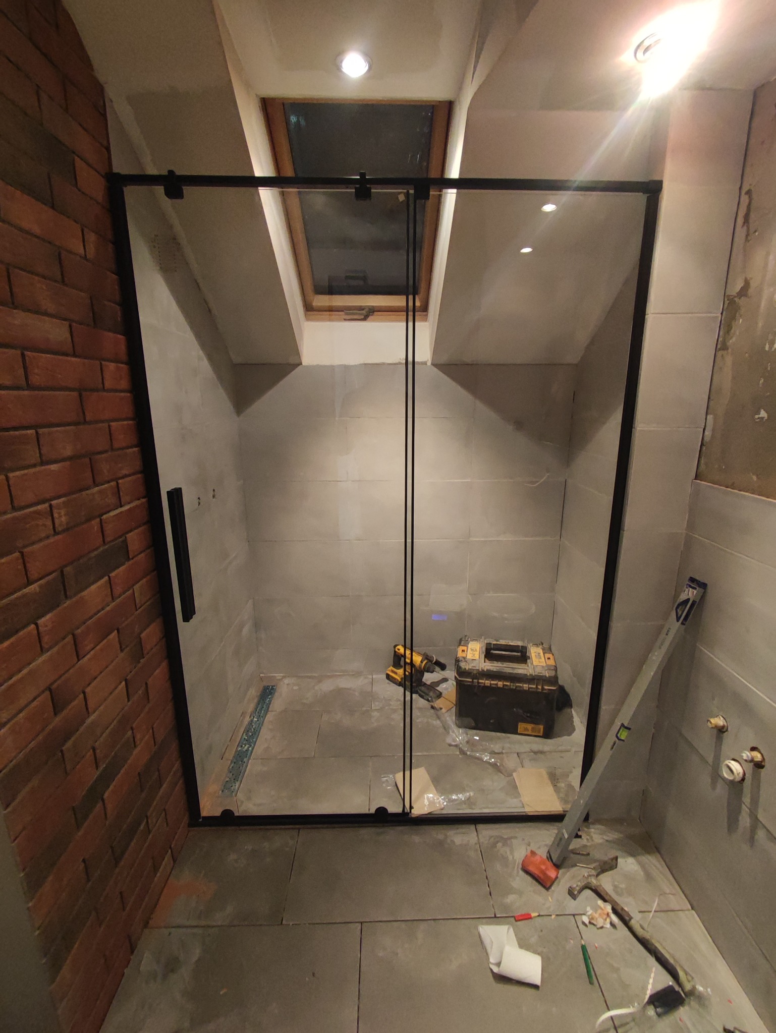 Montaż kabiny prysznicowej z czarnymi profilami w łazience z oknem dachowym, widoczne narzędzia i skrzynka narzędziowa, ściana z cegły i płytki w odcieniach szarości.