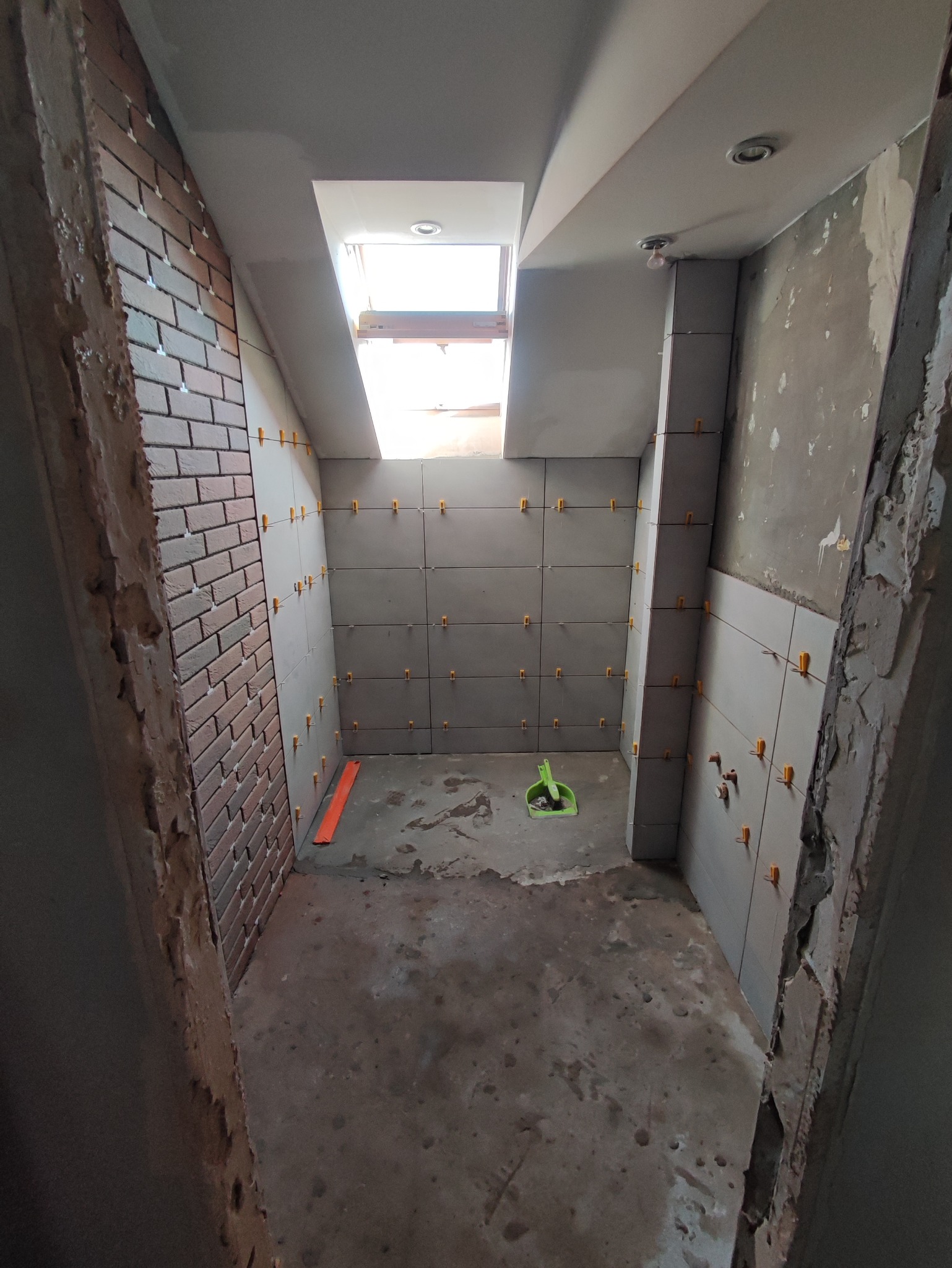 Łazienka w trakcie remontu: szare płytki z krzyżykami dystansowymi na ścianach, fragment ściany z cegły, okno dachowe, widoczny stan surowy podłogi i fragmenty ścian bez wykończenia.