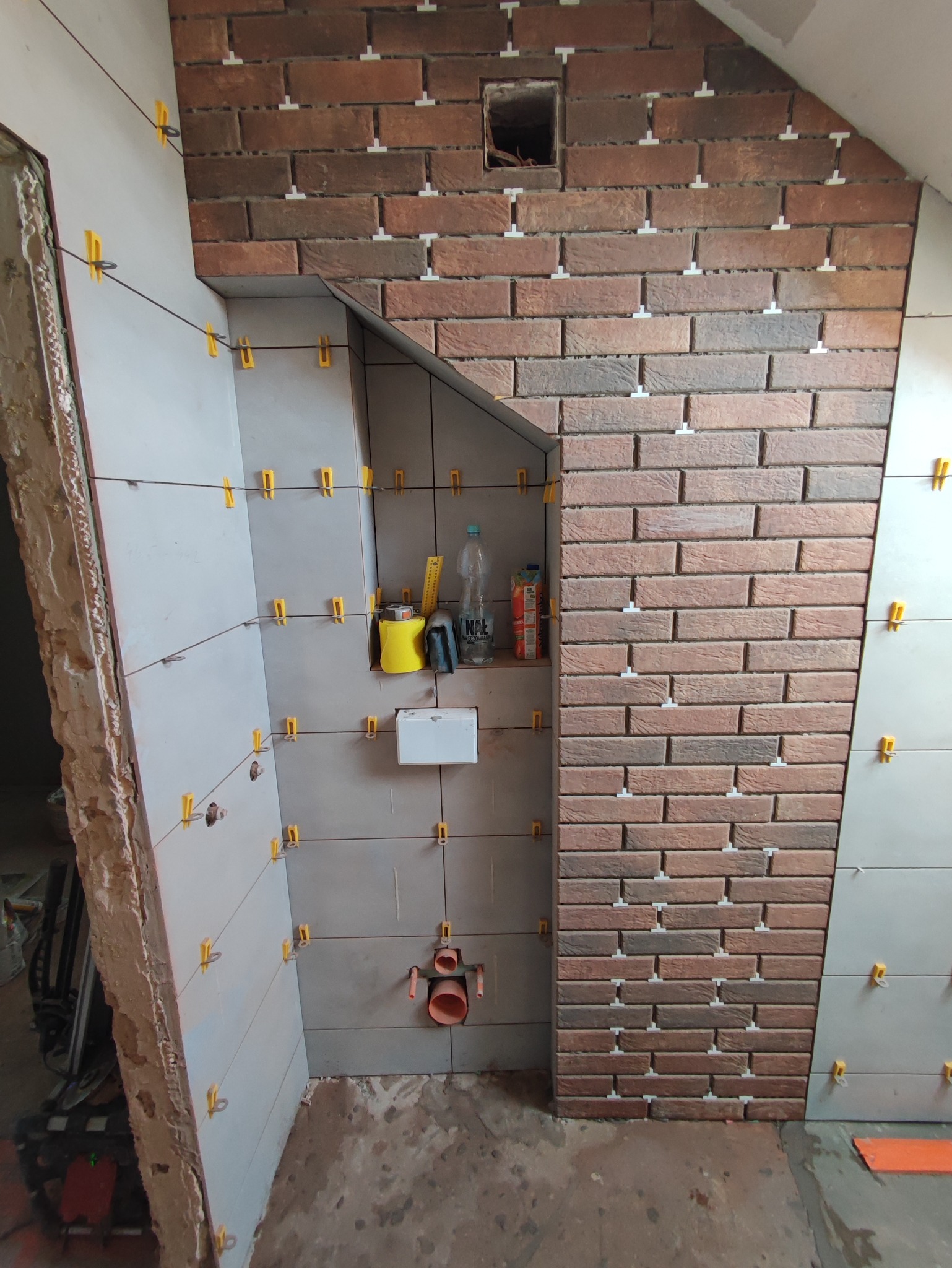 Ściana łazienki w trakcie układania płytek, widoczne płytki imitujące cegłę oraz szare płytki, użyte klipsy poziomujące, narzędzia i materiały na półce.