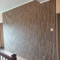 Ściana wykończona panelami pionowymi w kolorze drewna i ciemnego brązu, widoczny fragment podłogi z drewnopodobnych paneli, biały włącznik światła i gniazdko elektryczne, fragment balustrady schodów.