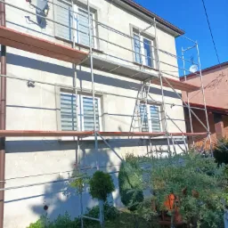 Dom jednorodzinny w trakcie prac elewacyjnych, widoczne rusztowania, błękitne niebo i fragment ogrodu z zielenią.