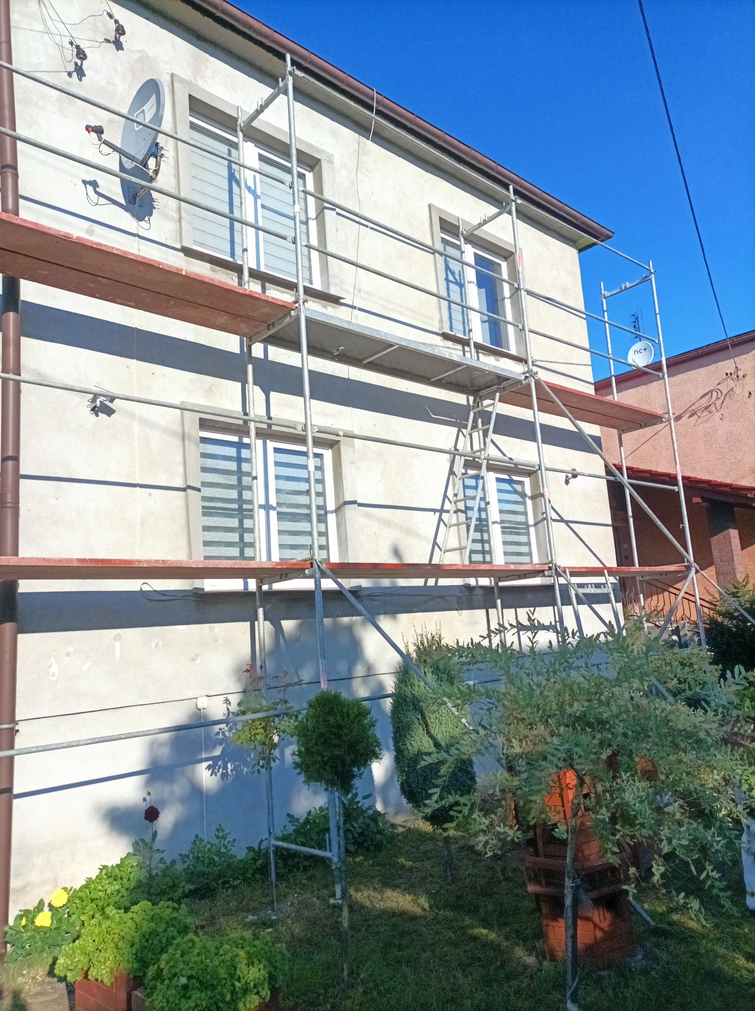 Dom jednorodzinny w trakcie prac elewacyjnych, widoczne rusztowania, błękitne niebo i fragment ogrodu z zielenią.