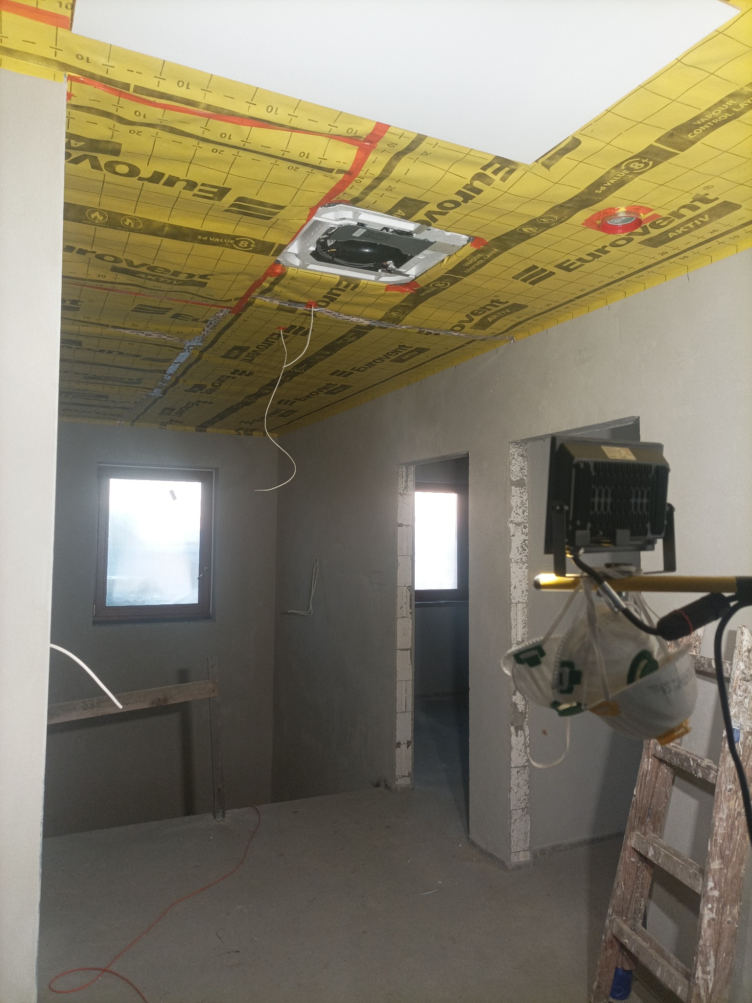 Poddasze w trakcie ocieplania, widoczna żółta folia Eurovent na suficie, surowe ściany, okno i drabina. Prace wykończeniowe w nowym domu.