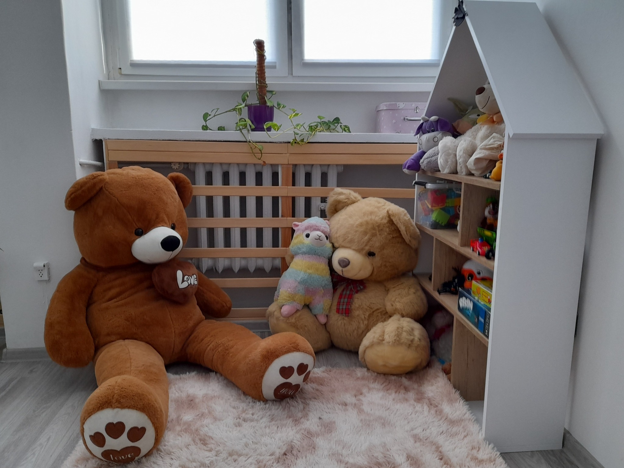 Pokój dziecięcy z dużym pluszowym misiem siedzącym na puszystym dywanie, obok mniejszy miś z alpakią, a w tle regał w kształcie domku wypełniony zabawkami.