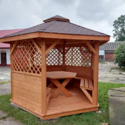 Drewniana altana ogrodowa z brązowym dachem krytym gontem bitumicznym, wyposażona w stół i ławki, na tle budynku z ludowymi motywami kwiatowymi.