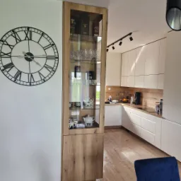 Nowoczesna kuchnia z białymi szafkami, drewnianym blatem i wysoką witryną z ekspozycją szkła i porcelany, uzupełniona dużym zegarem ściennym.