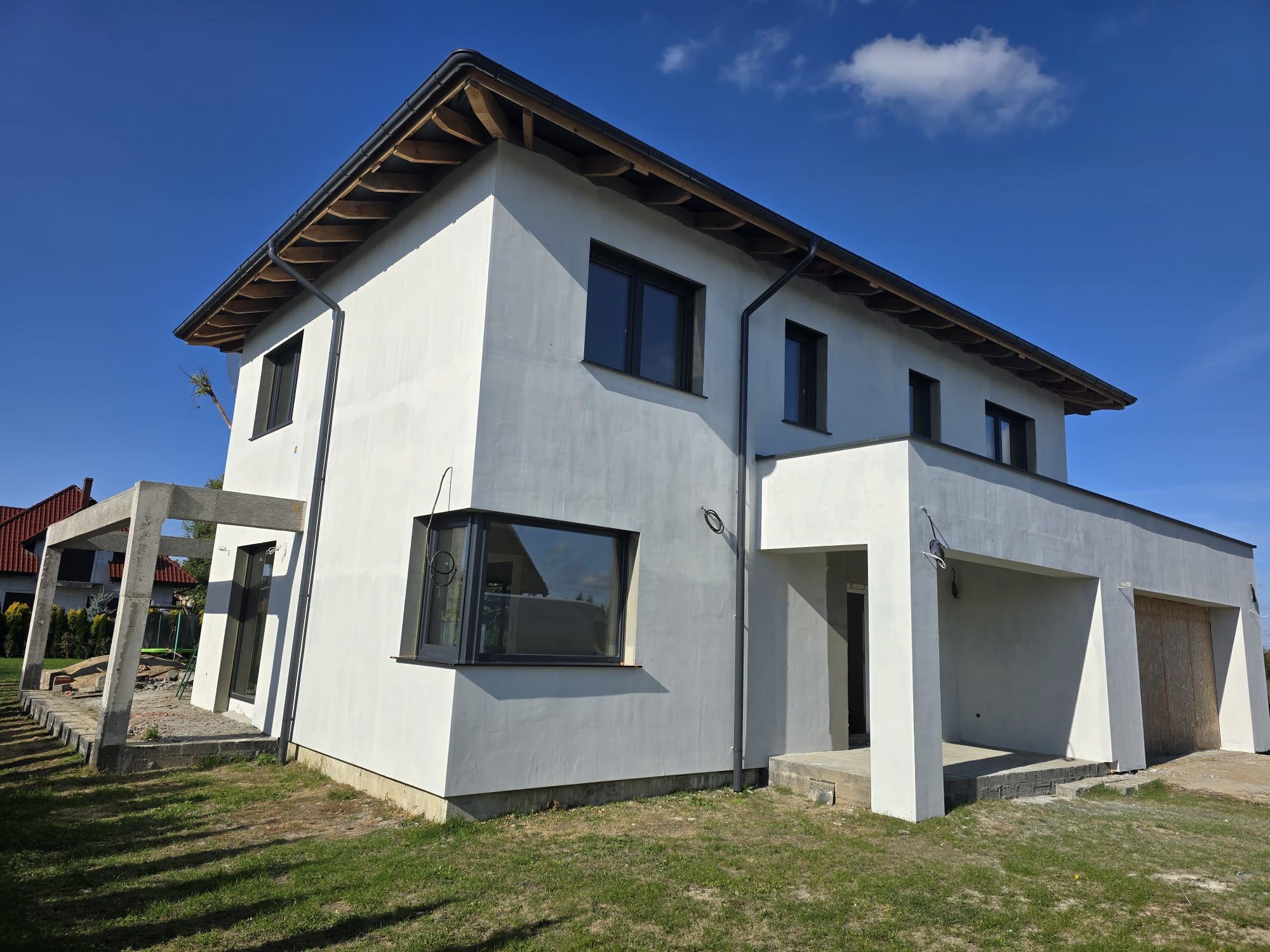 Nowoczesny dom jednorodzinny z jasną elewacją w trakcie prac wykończeniowych, widoczne elementy konstrukcyjne tarasu oraz fragmenty niezagospodarowanego terenu wokół budynku.