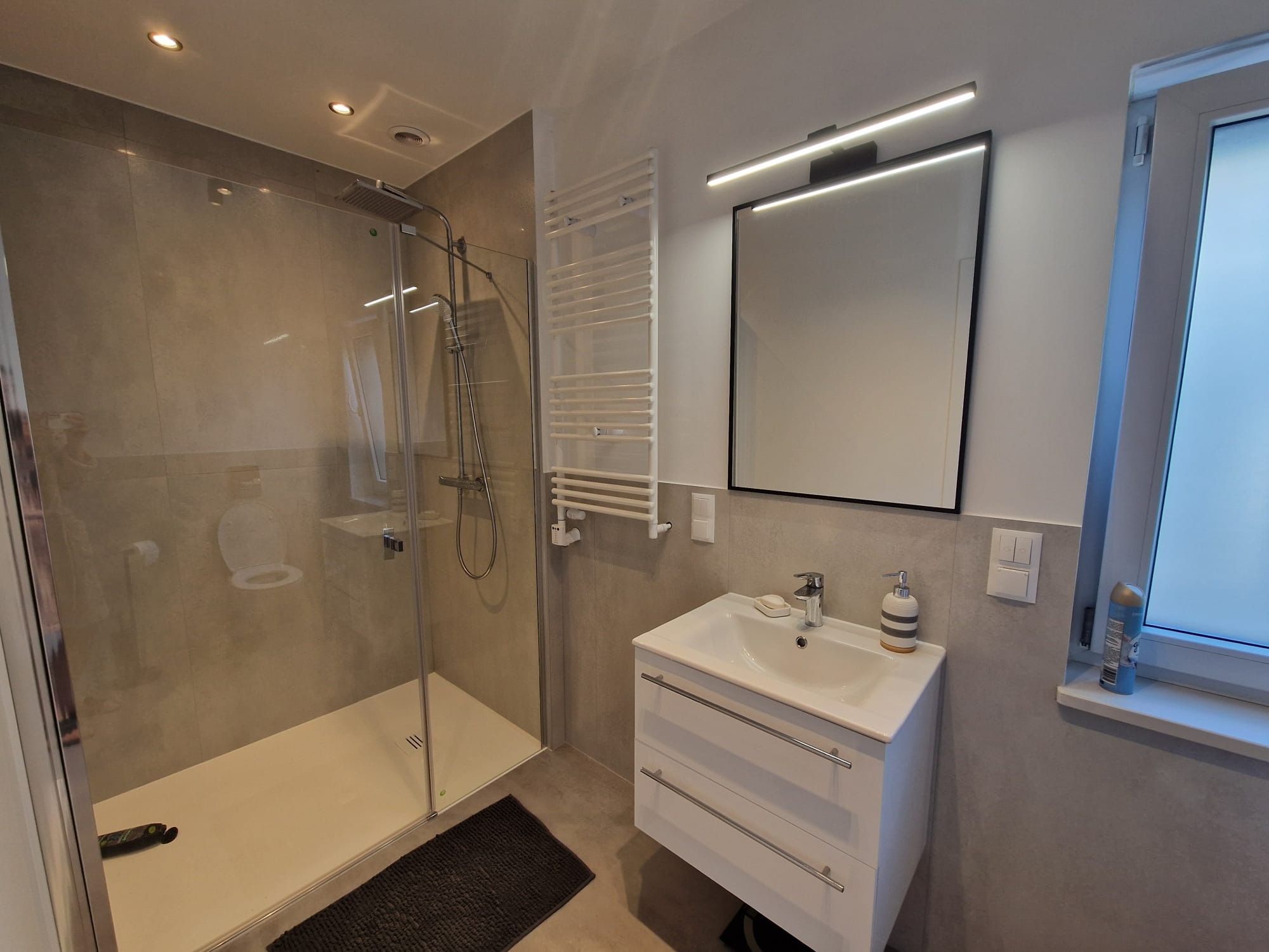 Nowoczesna łazienka z kabiną prysznicową typu walk-in, białą szafką umywalkową z dwoma szufladami i podświetlanym lustrem, grzejnik drabinkowy oraz szary dywanik pod prysznicem.