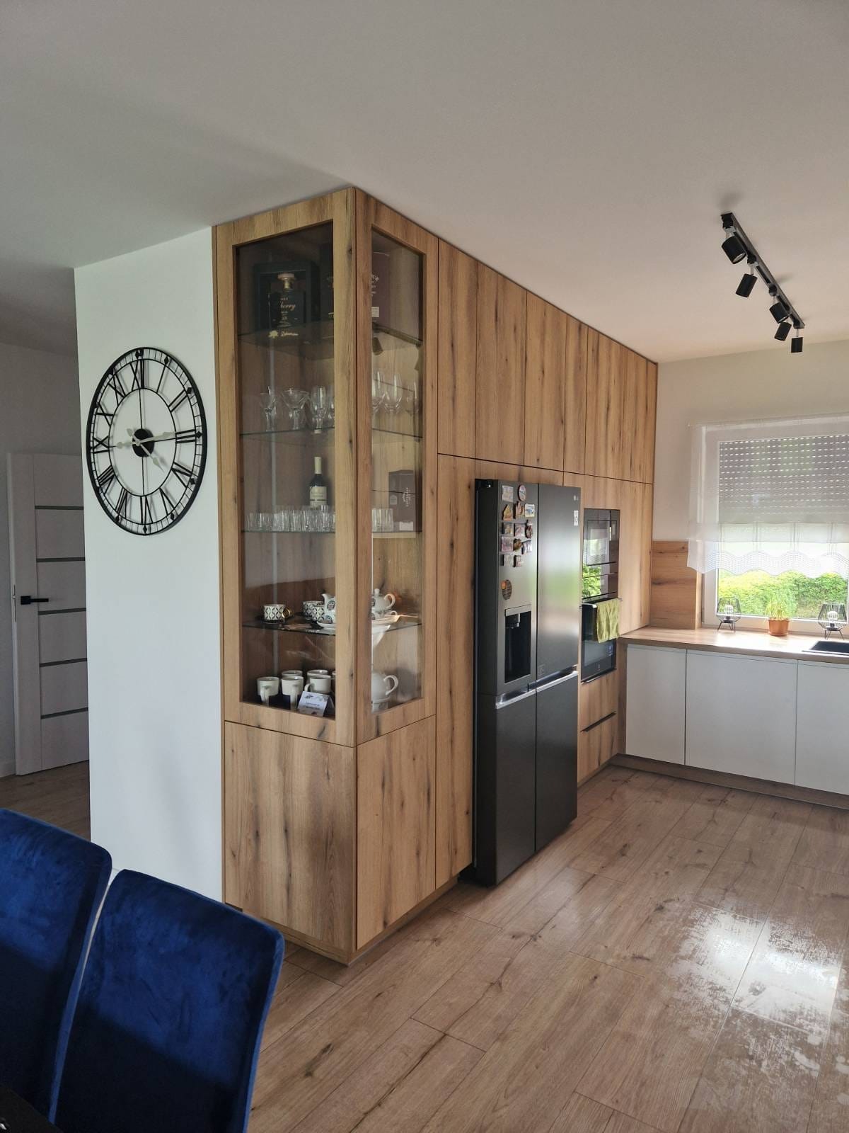 Nowoczesna kuchnia z drewnianymi szafkami, przeszkloną witryną z zastawą, czarną lodówką i zegarem ściennym w stylu vintage, oświetlona reflektorami sufitowymi.