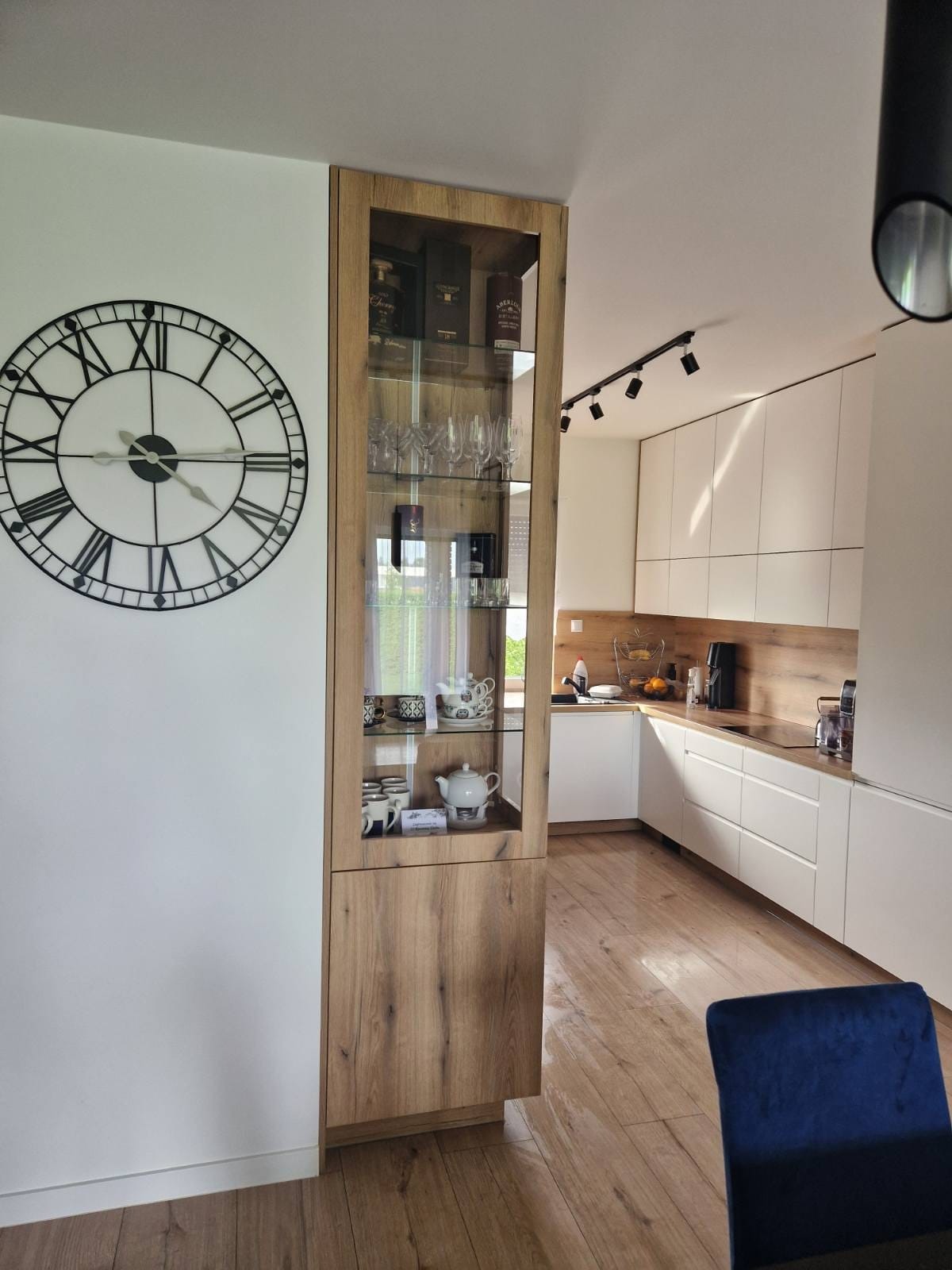 Nowoczesna kuchnia z białymi szafkami, drewnianym blatem i wysoką witryną z ekspozycją szkła i porcelany, uzupełniona dużym zegarem ściennym.