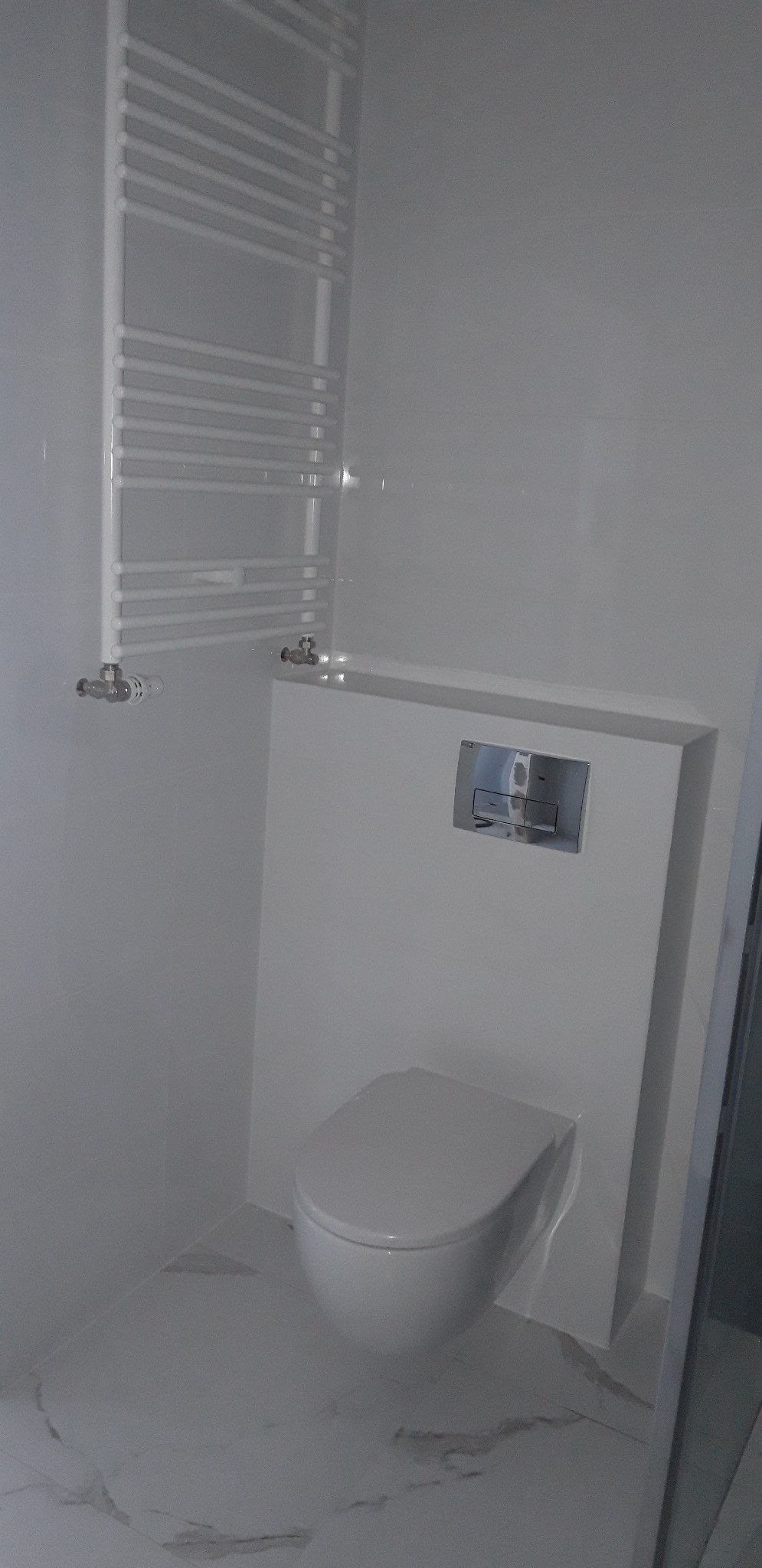 Biała, minimalistyczna łazienka z podwieszaną toaletą, chromowanym przyciskiem spłukiwania i grzejnikiem drabinkowym na ścianie z białych płytek.