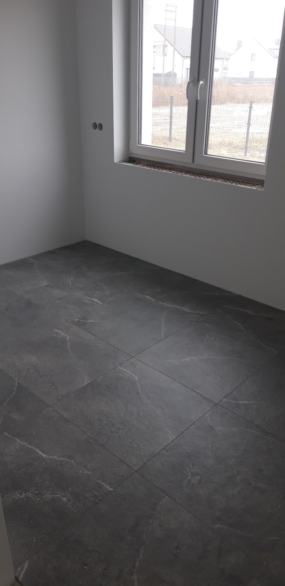 Ułożona, ciemnoszara podłoga z płytek imitujących kamień w pustym, jasnym pomieszczeniu z oknem, widoczne dwa otwory na gniazdka elektryczne na ścianie.