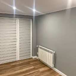 Szare pomieszczenie z drewnopodobną podłogą, białym grzejnikiem i roletami dzień-noc w oknach, świeżo po odnowieniu.