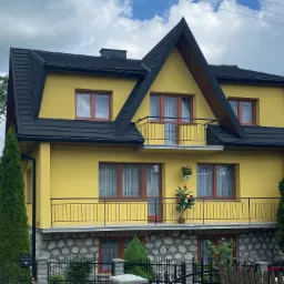 Żółty dom z czarnym dachem i balkonami, wykończony kamienną elewacją na parterze, z roślinami w doniczkach na balkonie.