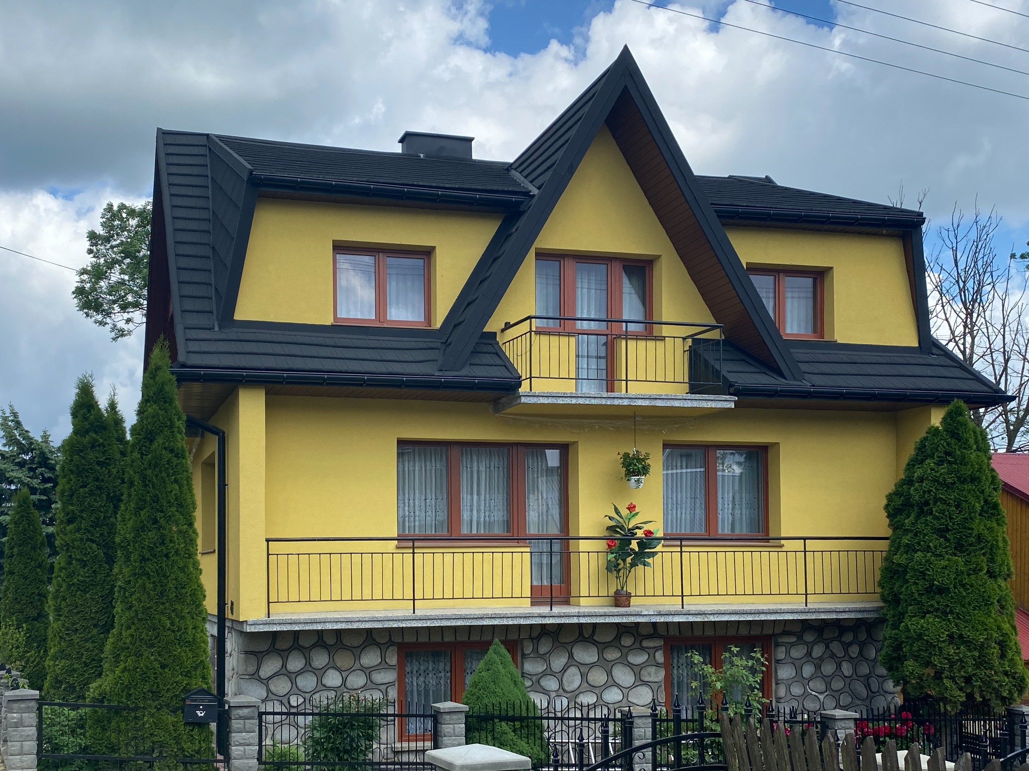 Żółty dom z czarnym dachem i balkonami, wykończony kamienną elewacją na parterze, z roślinami w doniczkach na balkonie.