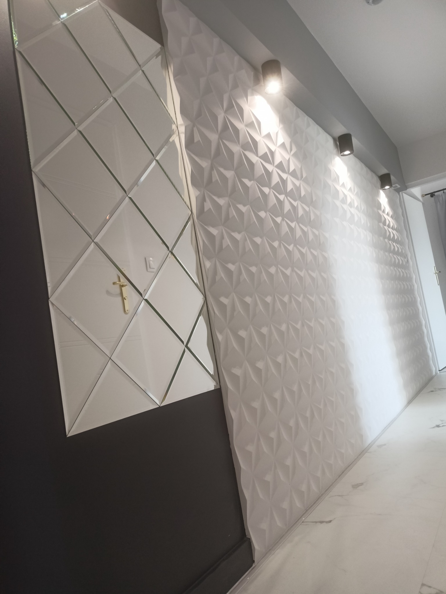Nowoczesna ściana z trójwymiarowych paneli gipsowych w kolorze białym, połączona z lustrzaną ścianą z geometrycznych płytek, oświetlona trzema punktowymi lampami sufitowymi, z widocznym fragmentem...