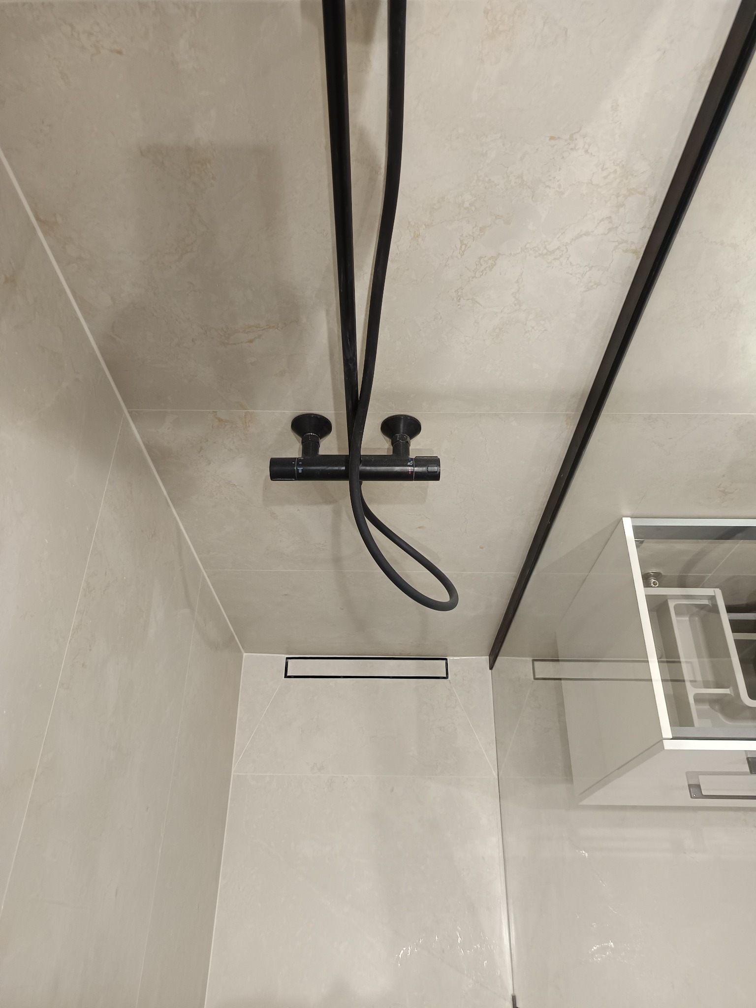 Beżowa łazienka z czarną armaturą: bateria prysznicowa i odpływ liniowy, widoczna kabina prysznicowa ze szklaną ścianą.