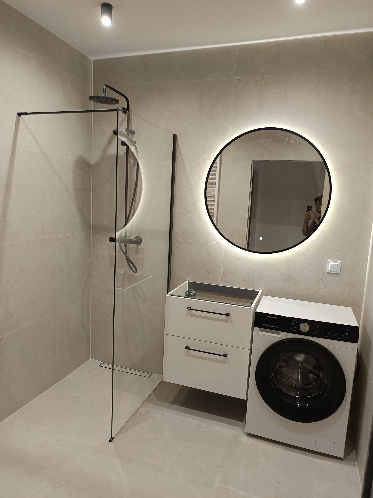Nowoczesna łazienka z kabiną prysznicową typu walk-in, okrągłym lustrem z podświetleniem LED, wiszącą szafką z szufladami i pralką Hisense, wykończona płytkami w odcieniach beżu.