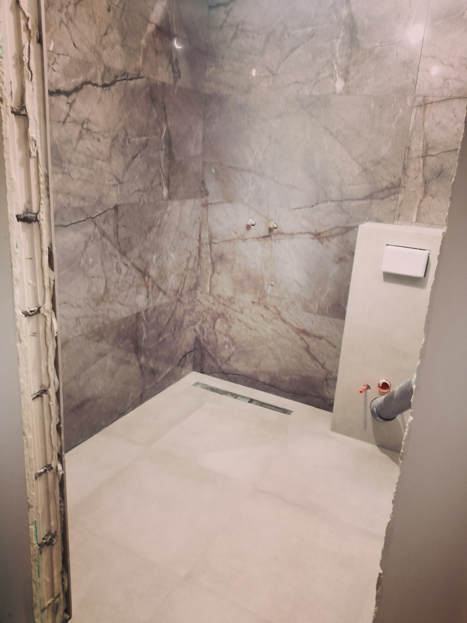 Wnętrze kabiny prysznicowej w trakcie prac wykończeniowych, ściany wyłożone płytami imitującymi marmur, widoczny odpływ liniowy, częściowo wykończona obudowa stelaża WC z instalacją hydrauliczną.