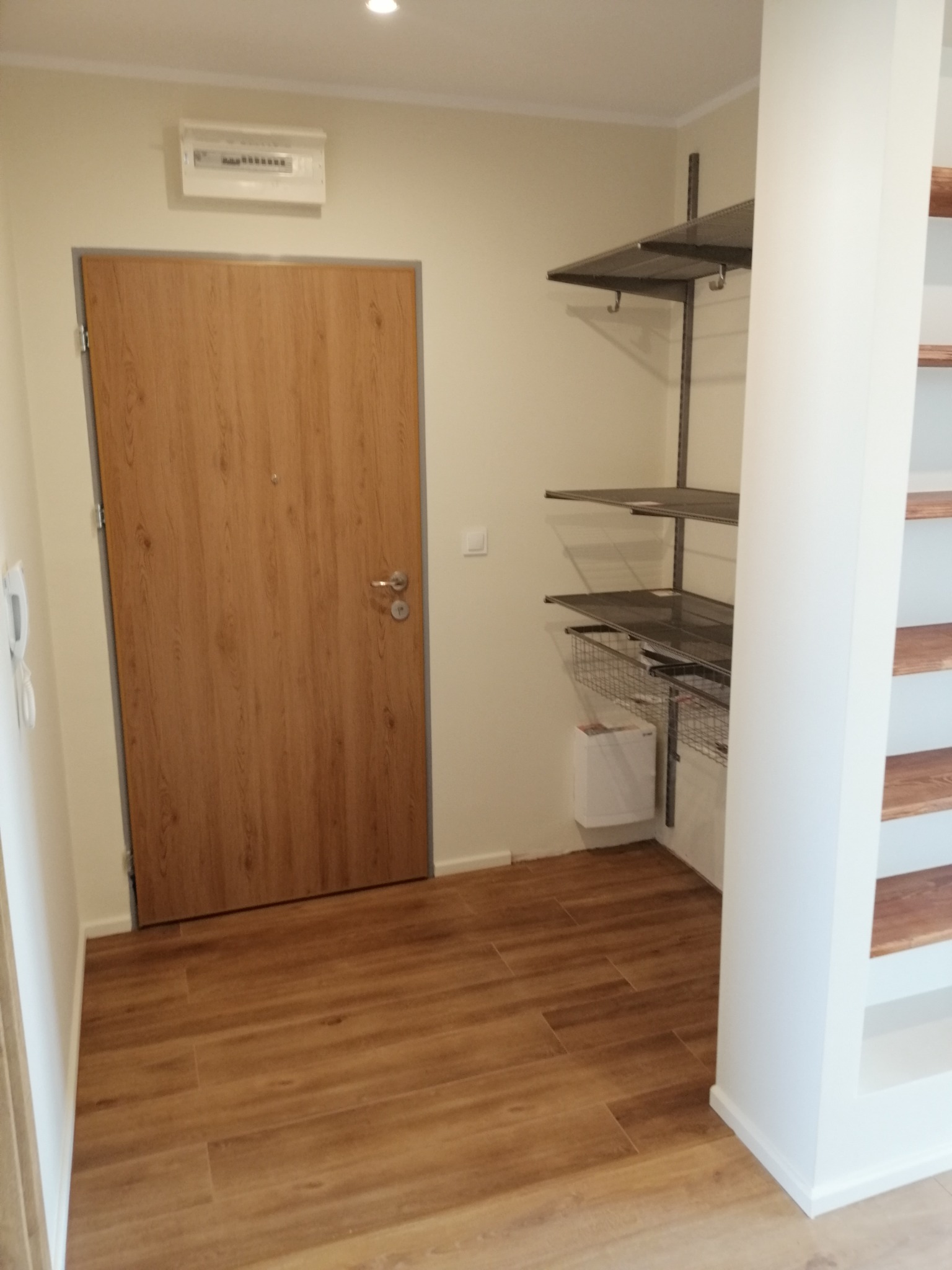 Przedpokój z drewnopodobną podłogą, drewnianymi drzwiami wejściowymi, metalowymi półkami na ścianie i zabudową regałową z drewnianymi półkami.