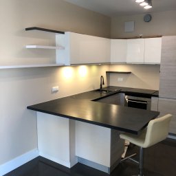 Glib Kolbasin - Nowoczesna kuchnia z czarnym blatem, białymi szafkami i ciemną podłogą, oświetlona punktowymi lampami sufitowymi, uzupełniona o hoker przy wyspie.