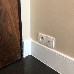Glib Kolbasin - Detal wykończenia wnętrza: połączenie ściany w odcieniu beżu z białą listwą przypodłogową, gniazdko elektryczne i gniazdo internetowe oraz fragment ciemnego, błyszczącego parkietu i drewnianych drzwi.