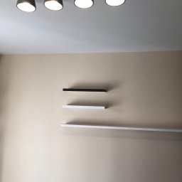 Glib Kolbasin - Beżowa ściana z trzema półkami (czarną, białą i długą białą) oraz sufit z lampą z pięcioma szarymi reflektorami na drewnianej listwie.