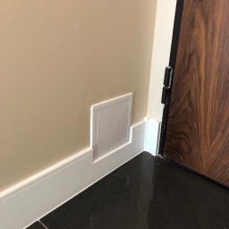 Glib Kolbasin - Ściana w odcieniach beżu z zamontowaną, ukrytą klapą rewizyjną w dolnej części, przy listwie przypodłogowej, obok ciemnych płytek podłogowych i brązowych drzwi.