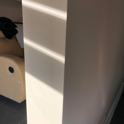 Glib Kolbasin - Fragment świeżo pomalowanej ściany w odcieniu beżu, oświetlonej promieniami słońca tworzącymi wyraźne pasy światła i cienia, widoczny narożnik ściany i fragment fotela w tle.