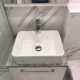 Glib Kolbasin - Biała, prostokątna umywalka nablatowa z chromowaną baterią na szafce z szufladami, blat z szarego granitu, tło z płytek imitujących marmur.