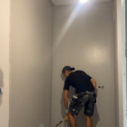 Glib Kolbasin - Mężczyzna w czarnej koszulce i wzorzystych spodenkach maluje ścianę w pomieszczeniu wałkiem, podłoga zabezpieczona folią malarską, widoczne puszki instalacyjne na ścianie.