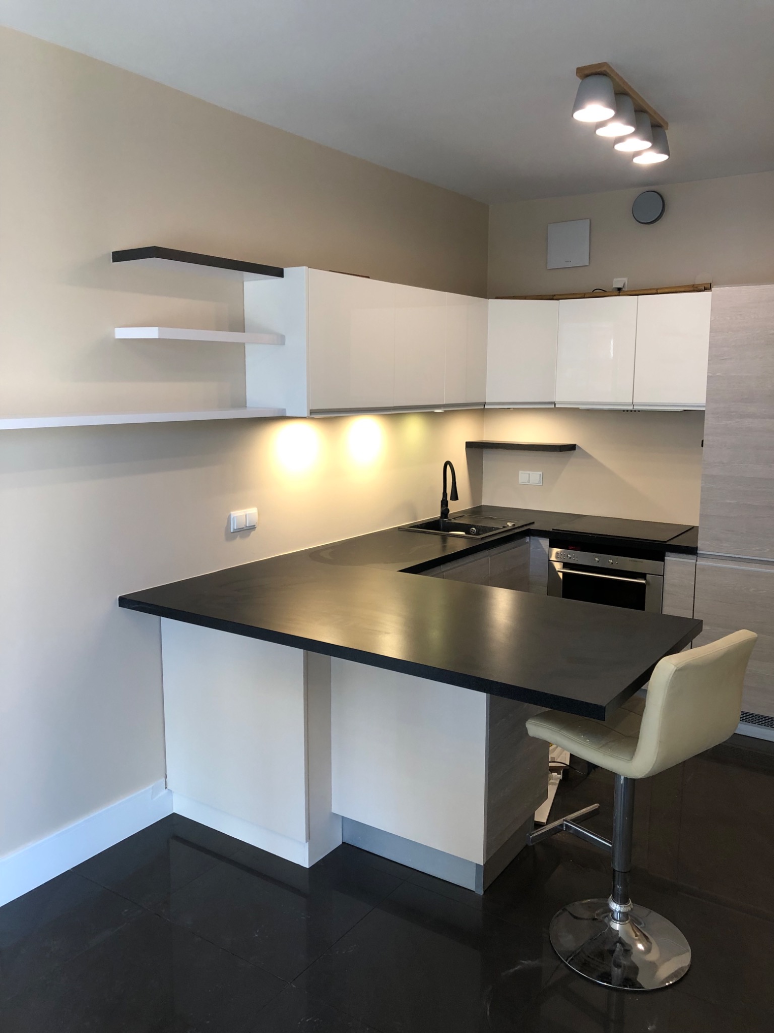 Nowoczesna kuchnia z czarnym blatem, białymi szafkami i ciemną podłogą, oświetlona punktowymi lampami sufitowymi, uzupełniona o hoker przy wyspie.