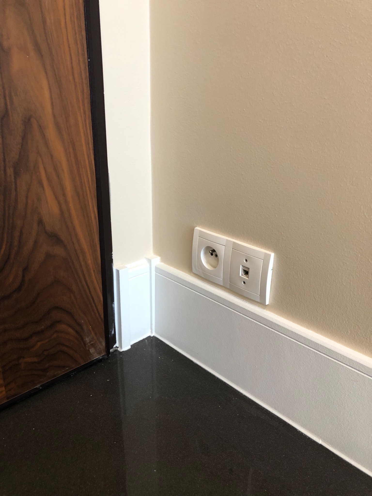 Detal wykończenia wnętrza: połączenie ściany w odcieniu beżu z białą listwą przypodłogową, gniazdko elektryczne i gniazdo internetowe oraz fragment ciemnego, błyszczącego parkietu i drewnianych drzwi.
