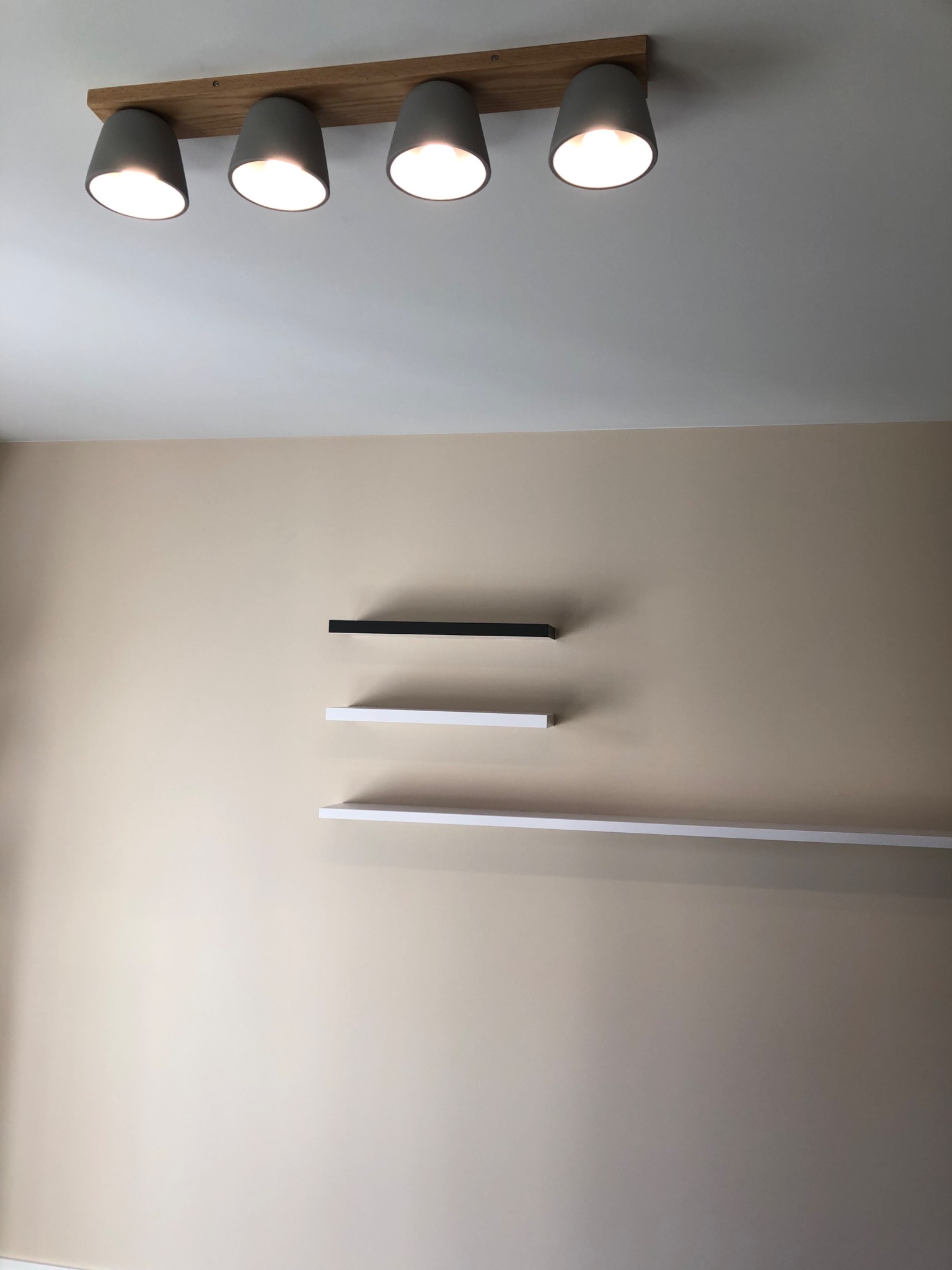 Beżowa ściana z trzema półkami (czarną, białą i długą białą) oraz sufit z lampą z pięcioma szarymi reflektorami na drewnianej listwie.