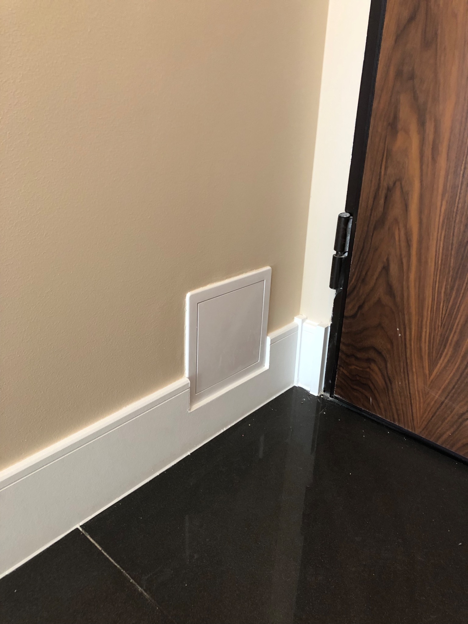 Ściana w odcieniach beżu z zamontowaną, ukrytą klapą rewizyjną w dolnej części, przy listwie przypodłogowej, obok ciemnych płytek podłogowych i brązowych drzwi.