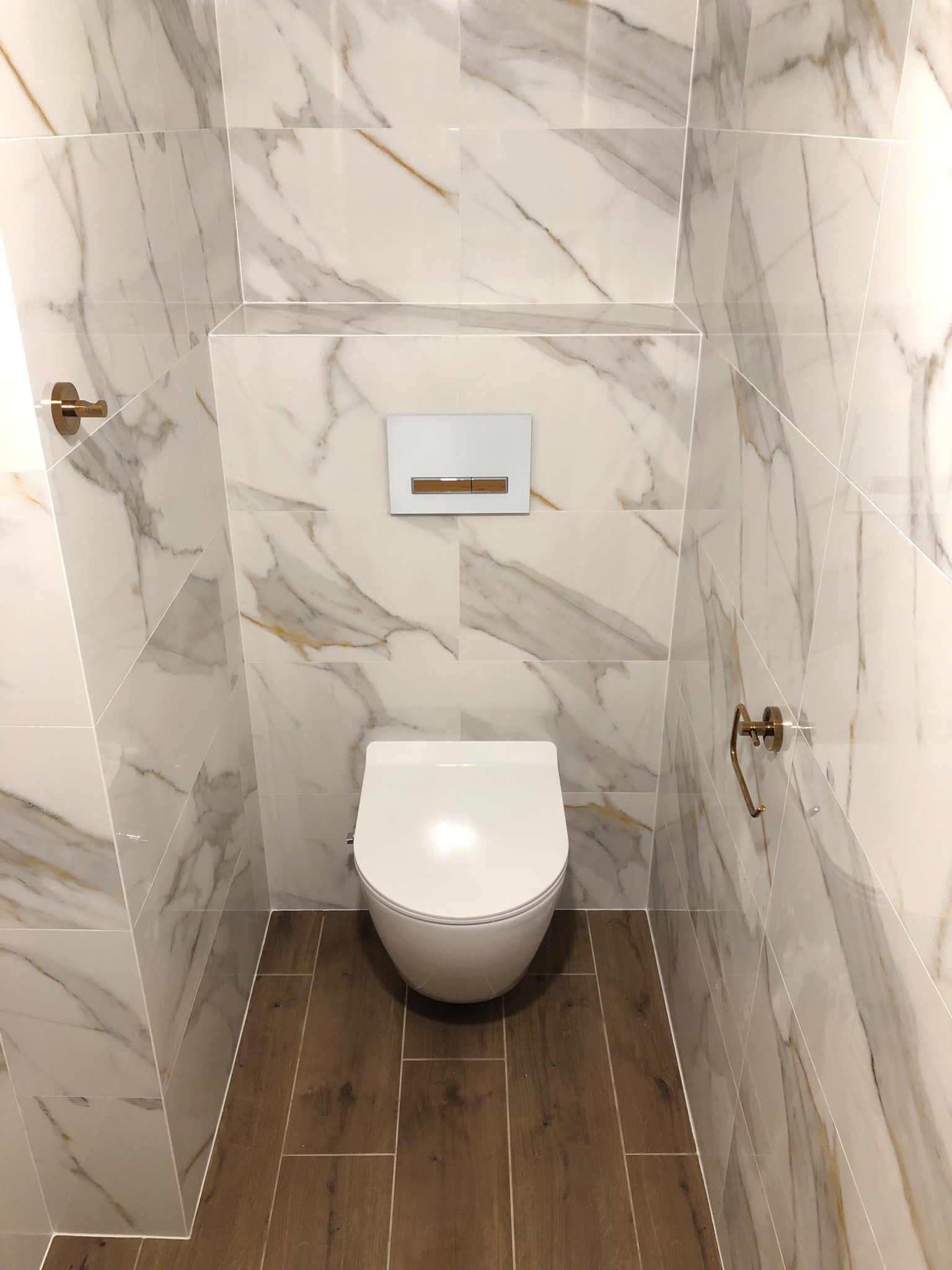Nowoczesna łazienka z podwieszaną toaletą, marmurowymi płytkami na ścianach z złotymi akcentami i podłogą imitującą drewno.