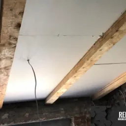 Płyty gipsowo-kartonowe zamontowane na drewnianych belkach stropowych, widoczne ślady po szpachlowaniu i wystający kabel elektryczny, fragment muru z cegły w tle.