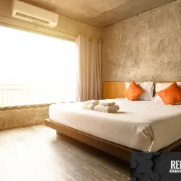 Nowoczesna sypialnia z surowym betonem na ścianie, jasnym oknem i minimalistycznym łóżkiem z pomarańczowymi poduszkami, widoczny klimatyzator i logo firmy w rogu kadru.